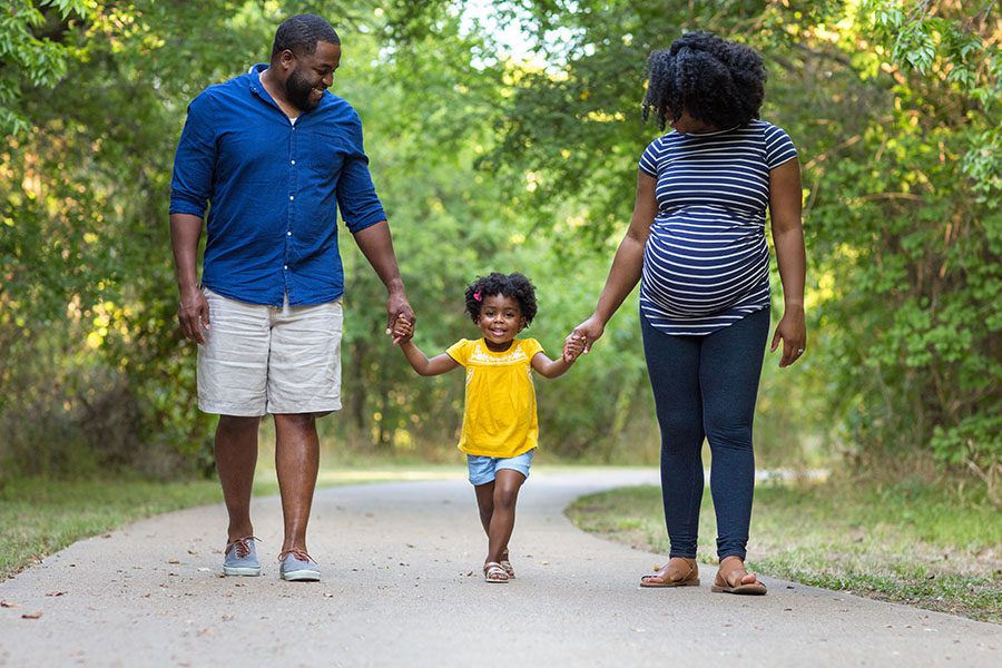 About Our Agency - Portrait of a Pregnant Woman Walking With Her Husband and Daughter on a Path at the Park on a Sunny Day