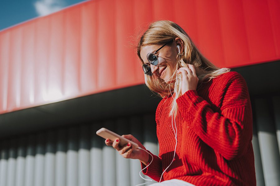Video Library - Side View Portrait of a Woman Wearing a Red Sweater With Earphones on Watching Videos on Her Smartphone and Smiling
