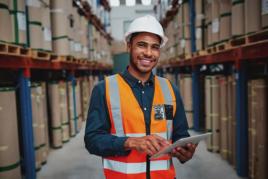 Warehousing and Logistics Insurance - Portrait of Smiling Warehouse Manager Using a Digital Tablet in a Warehouse While Standing between Shelves and Looking at the Camera with Helmet and Vest on